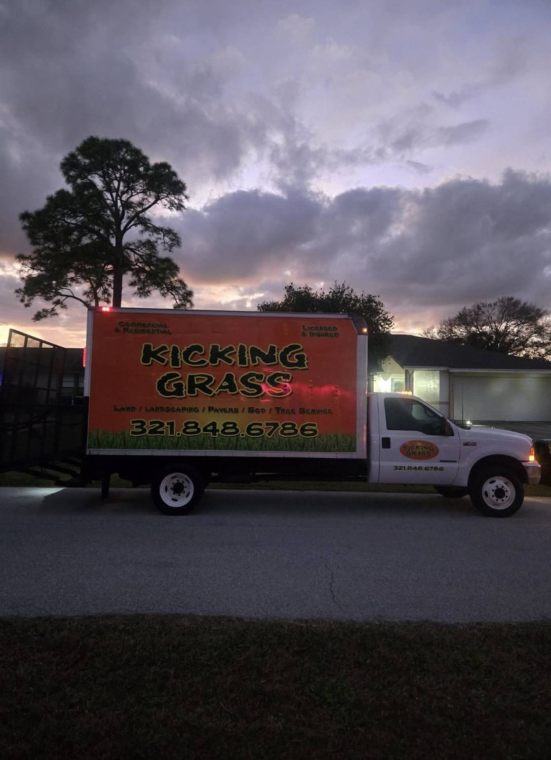 The orange KICKING GRASS box truck parked on a street at sunset