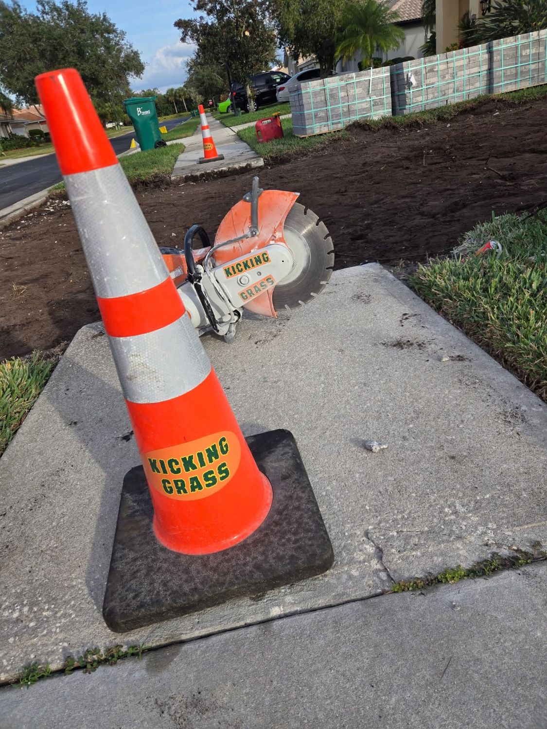 Orange traffic cone with a Kicking Grass sticker on a job site, next to a cut-off saw
