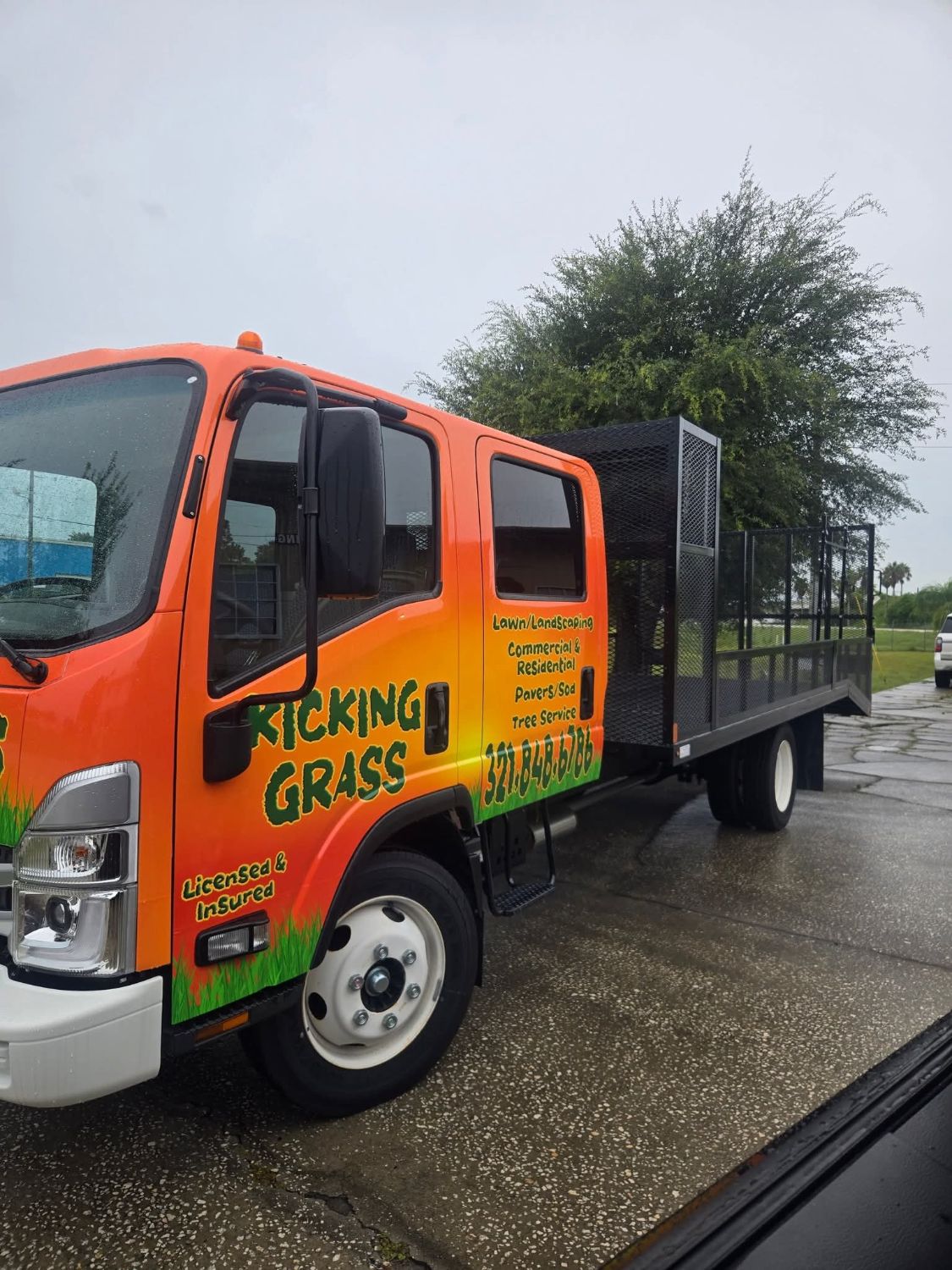 Orange Kicking Grass Isuzu landscape truck with a mesh cage bed, lettered Licensed and Insured