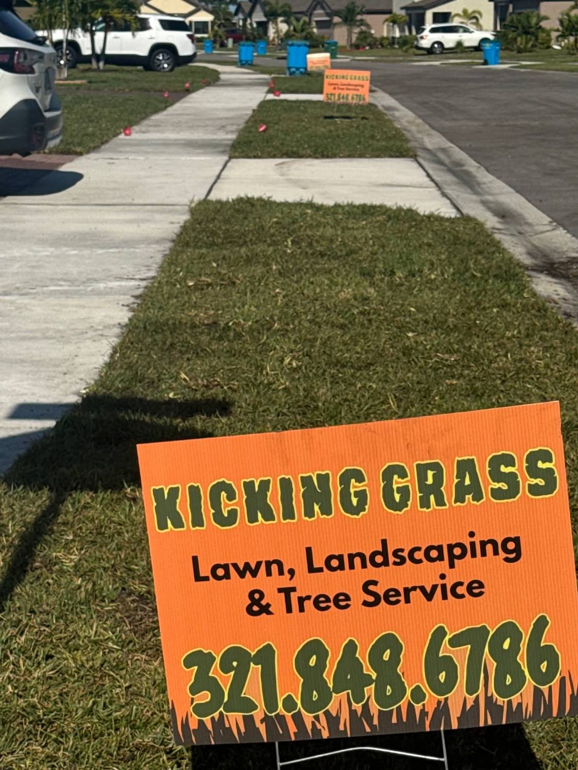 Orange Kicking Grass yard sign reading Lawn, Landscaping and Tree Service, 321.848.6786, on a fresh lawn